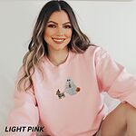Smiling woman in a Light Pink crewneck sweatshirt embroidered with a red dachshund in a skeleton costume and witch hat on a leash held by a ghost sipping iced coffee