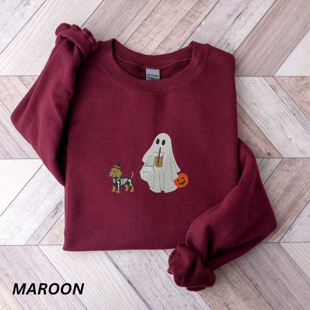 Dachshund Skeleton Sweatshirt – Maroon Maroon crewneck sweatshirt laid flat on a wood floor, embroidered with a red dachshund in a skeleton costume and witch hat on a leash held by a ghost sipping iced coffee