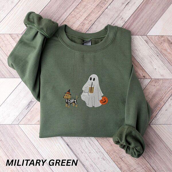 Military Green crewneck sweatshirt laid flat on a wood floor, embroidered with a red dachshund in a skeleton costume and witch hat on a leash held by a ghost sipping iced coffee