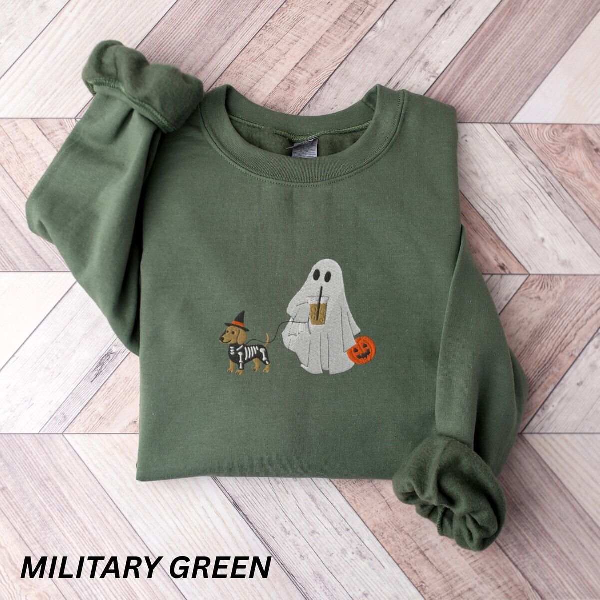Dachshund Skeleton Sweatshirt – Military Green Military Green crewneck sweatshirt laid flat on a wood floor, embroidered with a red dachshund in a skeleton costume and witch hat on a leash held by a ghost sipping iced coffee