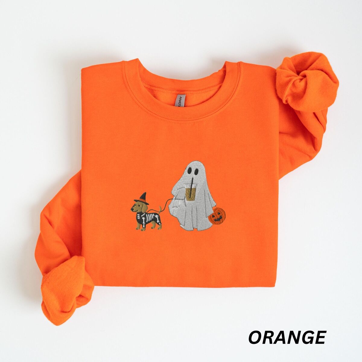 Dachshund Skeleton Sweatshirt - Orange Orange crewneck sweatshirt laid flat on white, embroidered with a red dachshund in a skeleton costume and witch hat on a leash held by a ghost sipping iced coffee