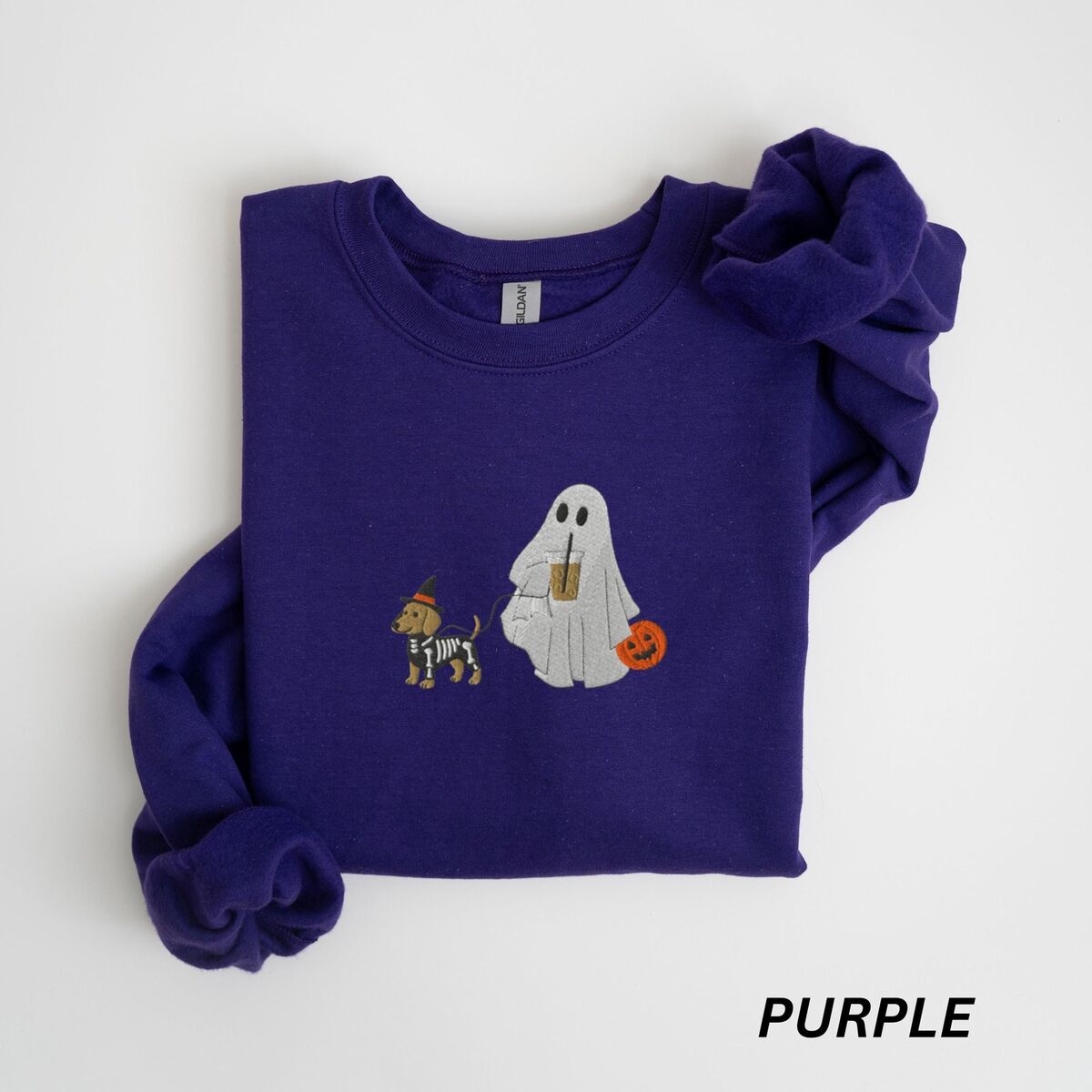 Dachshund Skeleton Sweatshirt - Purple Purple crewneck sweatshirt laid flat on white, embroidered with a red dachshund in a skeleton costume and witch hat on a leash held by a ghost sipping iced coffee