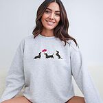 Smiling woman in a Ash crewneck sweatshirt embroidered with three black and tan dachshunds, the middle one carrying two pink heart balloons by the string