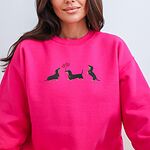Smiling woman in a Heliconia crewneck sweatshirt embroidered with three black and tan dachshunds, the middle one carrying two pink heart balloons by the string