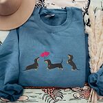 Indigo Blue crewneck sweatshirt laid flat with a tan hat and dried pampas, embroidered with three black and tan dachshunds, the middle one carrying two pink heart balloons by the string