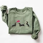 Military Green crewneck sweatshirt laid flat, embroidered with three black and tan dachshunds, the middle one carrying two pink heart balloons by the string