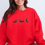 Smiling woman in a Red crewneck sweatshirt embroidered with three black and tan dachshunds, the middle one carrying two pink heart balloons by the string