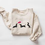 Sand crewneck sweatshirt laid flat, embroidered with three black and tan dachshunds, the middle one carrying two pink heart balloons by the string