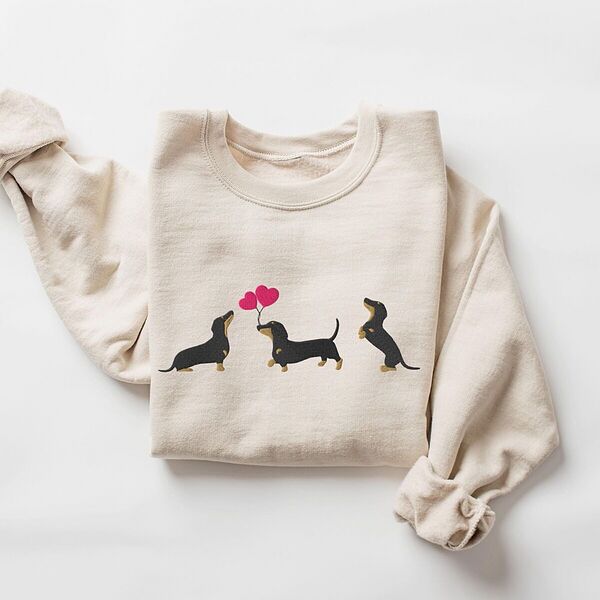 Sand crewneck sweatshirt laid flat, embroidered with three black and tan dachshunds, the middle one carrying two pink heart balloons by the string
