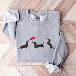 Sport Grey crewneck sweatshirt laid flat on a wood floor, embroidered with three black and tan dachshunds, the middle one carrying two pink heart balloons by the string