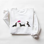 White crewneck sweatshirt laid flat, embroidered with three black and tan dachshunds, the middle one carrying two pink heart balloons by the string