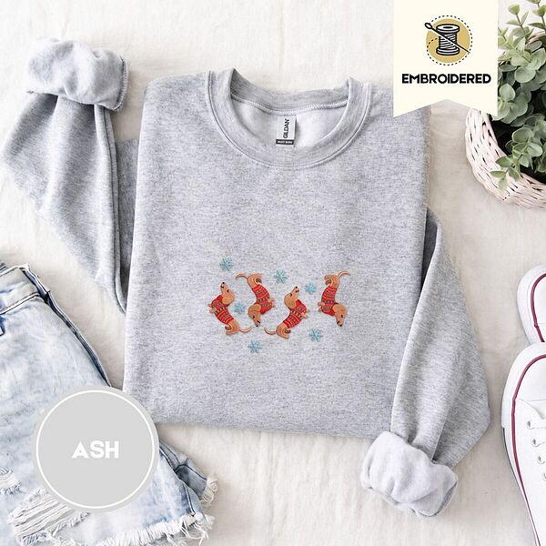 Ash crewneck sweatshirt folded with denim shorts, white sneakers and a potted plant, embroidered with four red dachshunds in red Fair Isle sweaters tumbling among light blue snowflakes