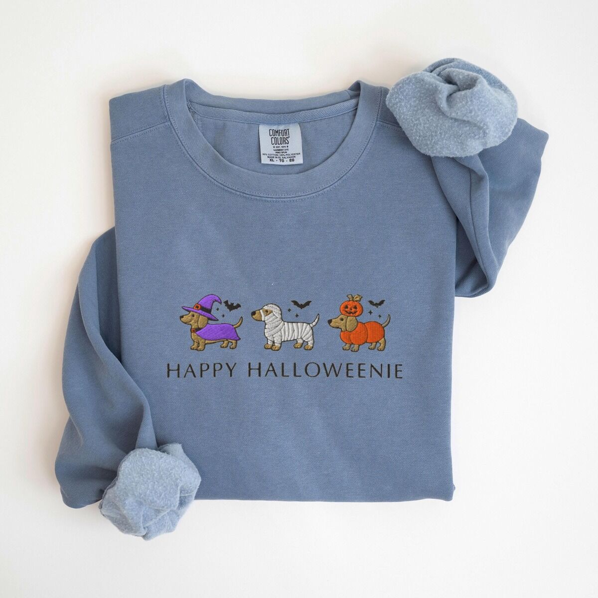 Dachshunds in Halloween Costumes Sweatshirt – Blue Jean Blue Jean crewneck sweatshirt laid flat on white, embroidered with three dachshunds dressed as a witch, a mummy and a pumpkin with bats overhead, above the words HAPPY HALLOWEENIE
