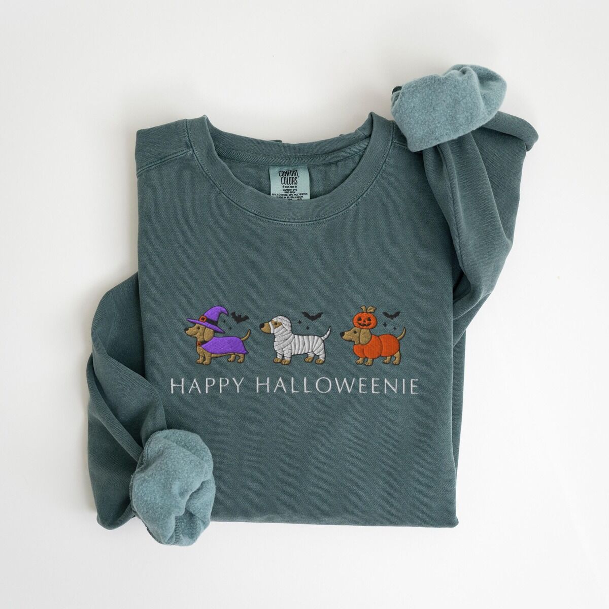 Dachshunds in Halloween Costumes Sweatshirt – Blue Spruce Blue Spruce crewneck sweatshirt laid flat on white, embroidered with three dachshunds dressed as a witch, a mummy and a pumpkin with bats overhead, above the words HAPPY HALLOWEENIE