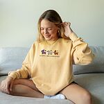 Woman sitting on a sofa in a Butter crewneck sweatshirt embroidered with three dachshunds dressed as a witch, a mummy and a pumpkin with bats overhead, above the words HAPPY HALLOWEENIE