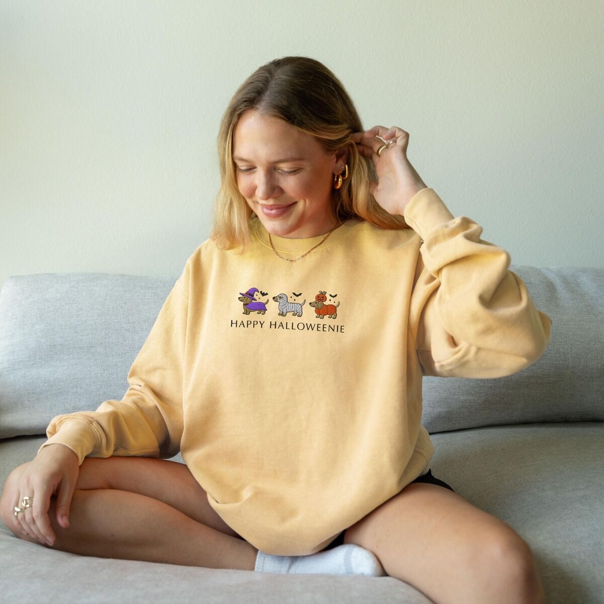 Dachshunds in Halloween Costumes Sweatshirt – Butter Woman sitting on a sofa in a Butter crewneck sweatshirt embroidered with three dachshunds dressed as a witch, a mummy and a pumpkin with bats overhead, above the words HAPPY HALLOWEENIE