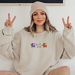 Smiling woman in a beanie wearing a Sand crewneck sweatshirt embroidered with three dachshunds dressed as a witch, a mummy and a pumpkin with bats overhead, no words