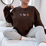 Woman sitting on a bed in a Dark Chocolate crewneck sweatshirt embroidered with three dachshunds dressed as a witch, a mummy and a pumpkin with bats overhead