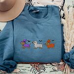 Indigo Blue crewneck sweatshirt laid flat with a straw hat, embroidered with three dachshunds dressed as a witch, a mummy and a pumpkin with bats overhead