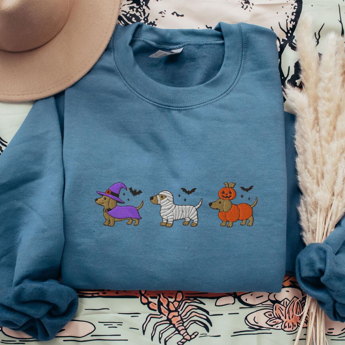 Dachshunds in Halloween Costumes Sweatshirt - Indigo Blue Indigo Blue crewneck sweatshirt laid flat with a straw hat, embroidered with three dachshunds dressed as a witch, a mummy and a pumpkin with bats overhead