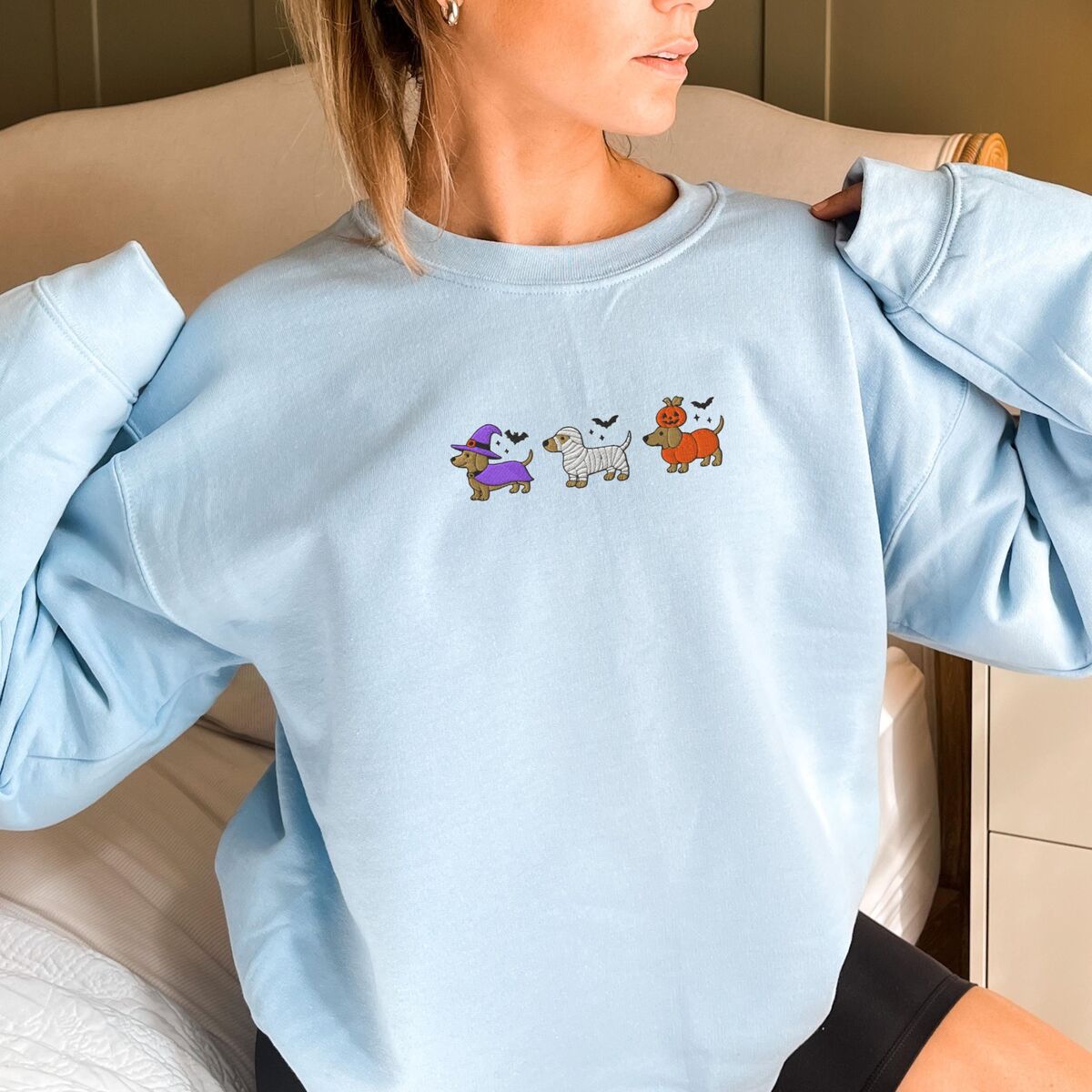 Dachshunds in Halloween Costumes Sweatshirt - Light Blue Woman in a Light Blue crewneck sweatshirt embroidered with three dachshunds dressed as a witch, a mummy and a pumpkin with bats overhead