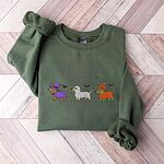 Military Green crewneck sweatshirt laid flat on a wood floor, embroidered with three dachshunds dressed as a witch, a mummy and a pumpkin with bats overhead