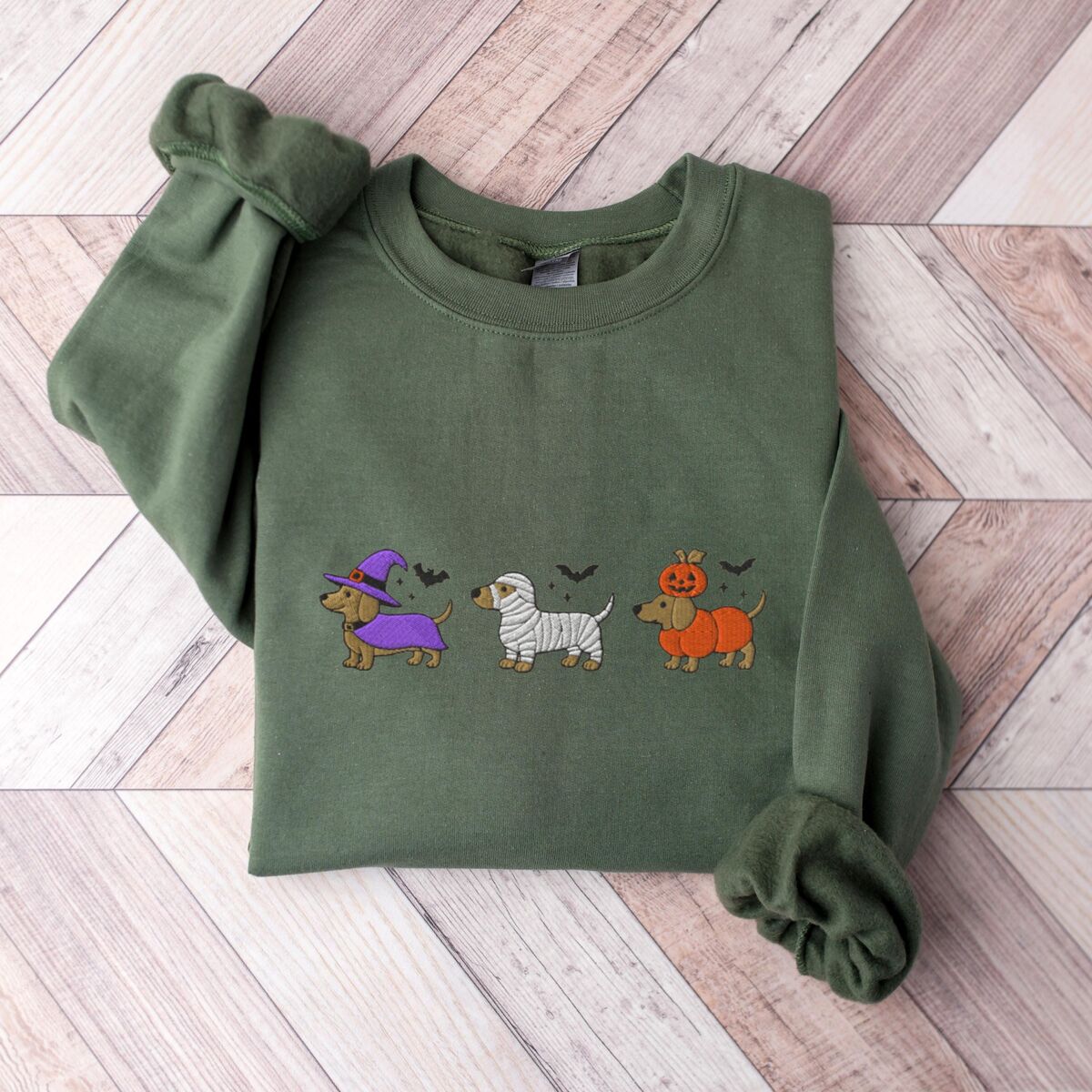 Dachshunds in Halloween Costumes Sweatshirt - Military Green Military Green crewneck sweatshirt laid flat on a wood floor, embroidered with three dachshunds dressed as a witch, a mummy and a pumpkin with bats overhead