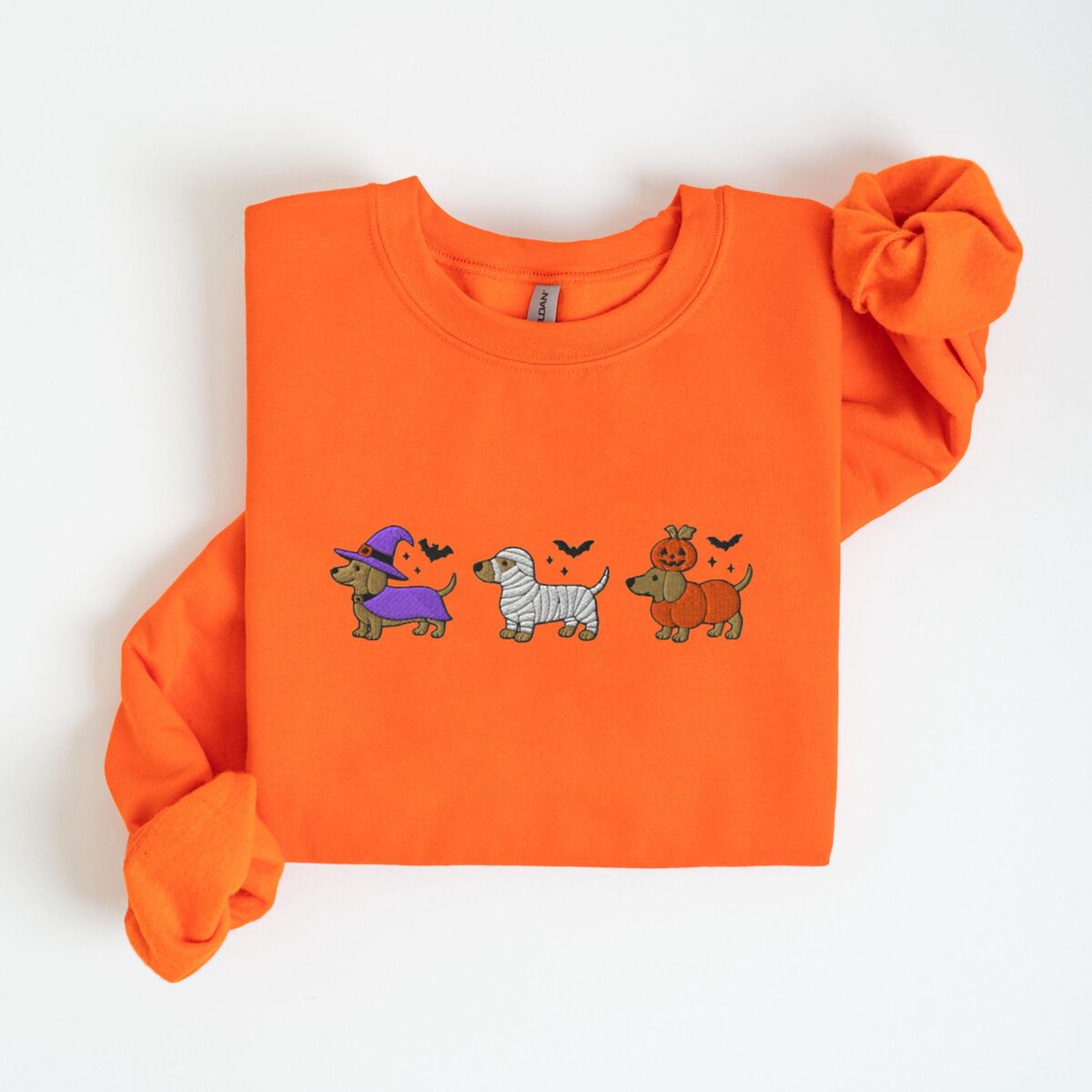 Dachshunds in Halloween Costumes Sweatshirt - Orange Orange crewneck sweatshirt laid flat on white, embroidered with three dachshunds dressed as a witch, a mummy and a pumpkin with bats overhead