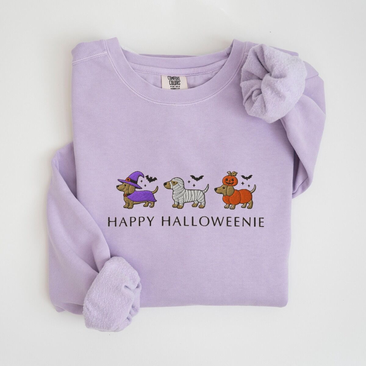 Dachshunds in Halloween Costumes Sweatshirt – Orchid Orchid crewneck sweatshirt laid flat on white, embroidered with three dachshunds dressed as a witch, a mummy and a pumpkin with bats overhead, above the words HAPPY HALLOWEENIE