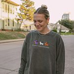 Woman on a tree-lined street in a Pepper crewneck sweatshirt embroidered with three dachshunds dressed as a witch, a mummy and a pumpkin with bats overhead, above the words HAPPY HALLOWEENIE