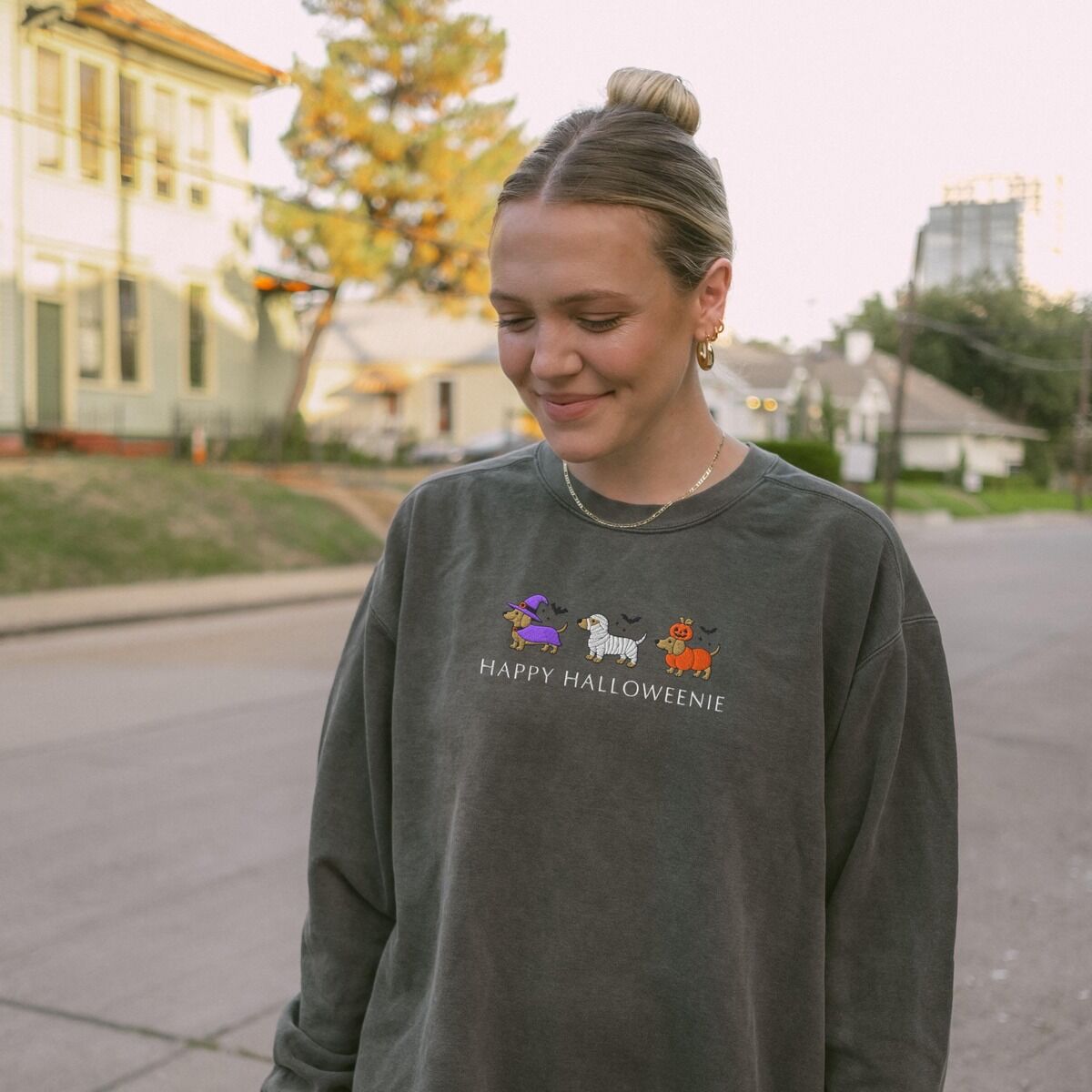 Dachshunds in Halloween Costumes Sweatshirt – Pepper Woman on a tree-lined street in a Pepper crewneck sweatshirt embroidered with three dachshunds dressed as a witch, a mummy and a pumpkin with bats overhead, above the words HAPPY HALLOWEENIE