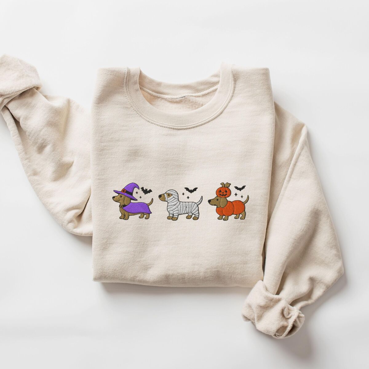 Dachshunds in Halloween Costumes Sweatshirt - Sand Sand crewneck sweatshirt laid flat on white, embroidered with three dachshunds dressed as a witch, a mummy and a pumpkin with bats overhead