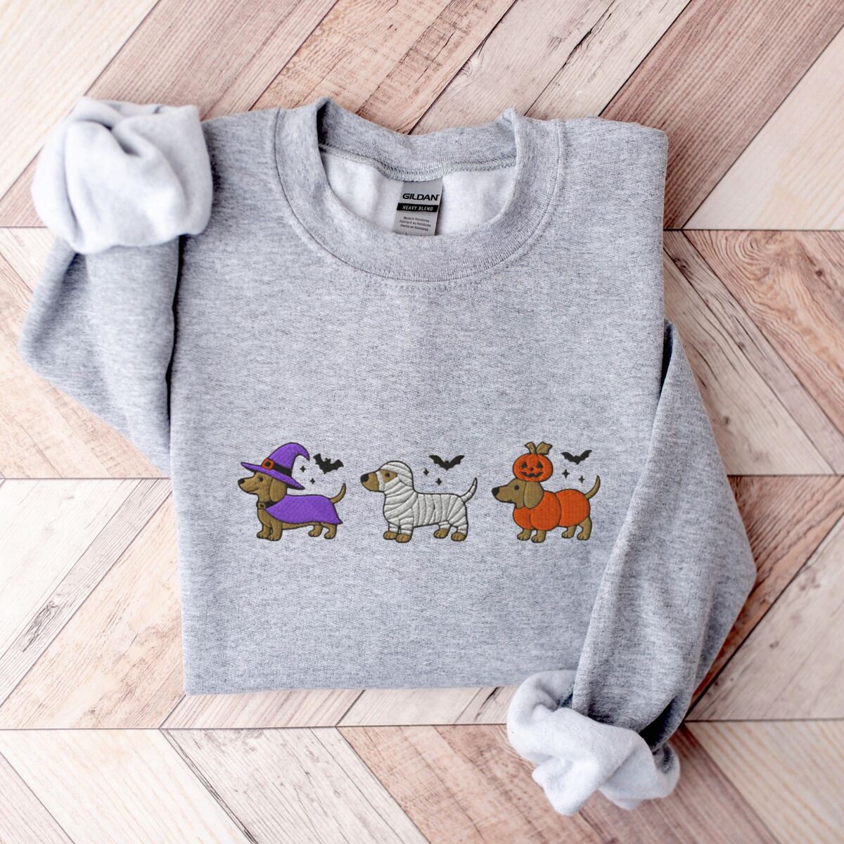 Dachshunds in Halloween Costumes Sweatshirt - Sport Grey Sport Grey crewneck sweatshirt laid flat on a wood floor, embroidered with three dachshunds dressed as a witch, a mummy and a pumpkin with bats overhead