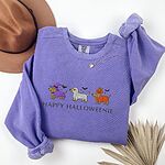 Violet crewneck sweatshirt laid flat with a hat and dried palm leaf, embroidered with three dachshunds dressed as a witch, a mummy and a pumpkin with bats overhead, above the words HAPPY HALLOWEENIE