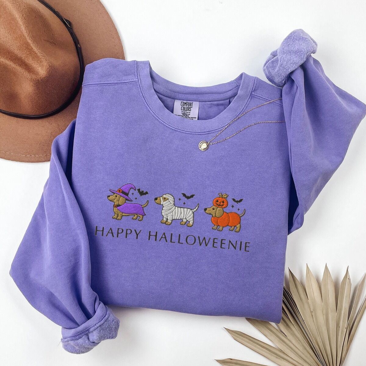 Dachshunds in Halloween Costumes Sweatshirt – Violet Violet crewneck sweatshirt laid flat with a hat and dried palm leaf, embroidered with three dachshunds dressed as a witch, a mummy and a pumpkin with bats overhead, above the words HAPPY HALLOWEENIE