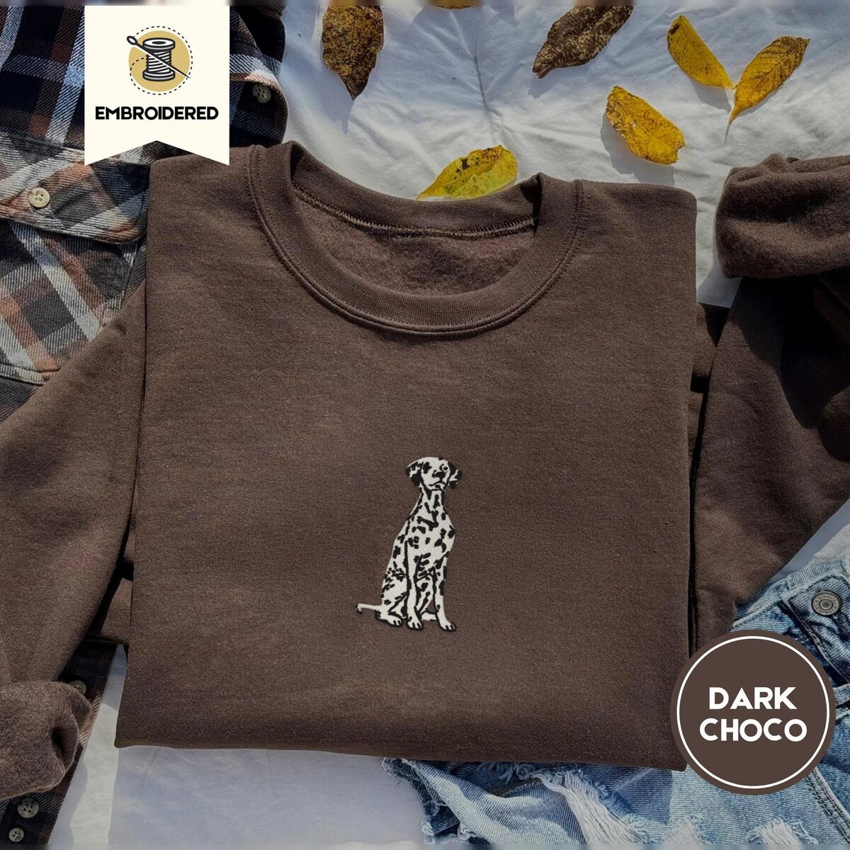 Dalmatian Sweatshirt – Dark Chocolate Dark Chocolate crewneck sweatshirt on a plaid flannel shirt with yellow autumn leaves and denim shorts, embroidered with a sitting Dalmatian in white with black spots and a black outline