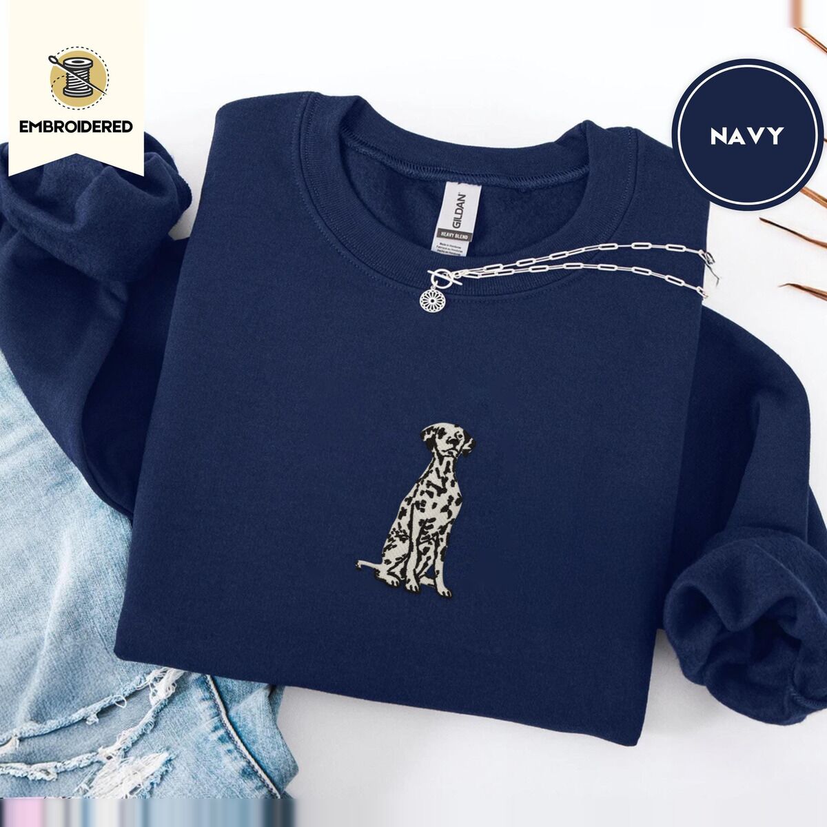 Dalmatian Sweatshirt – Navy Navy crewneck sweatshirt folded with ripped jeans and a silver chain necklace, embroidered with a sitting Dalmatian in white with black spots and a black outline