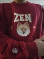 Man wearing a maroon sweatshirt embroidered with a Pomeranian portrait, the name ZEN, EST year and a white paw print on the sleeve