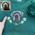 Forest green dog memorial sweatshirt with an embroidered gray dog portrait, a white daisy wreath and the name Gus in white script, beside the original photo