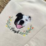 Close-up of an embroidered black and white dog face with a yellow and orange wildflower wreath and the name Mangchi in black script on a sand sweatshirt