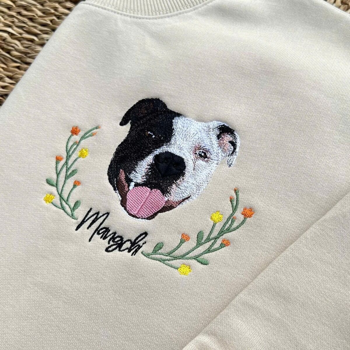 Dog Memorial Sweatshirt – Wildflower Wreath Close-up Close-up of an embroidered black and white dog face with a yellow and orange wildflower wreath and the name Mangchi in black script on a sand sweatshirt