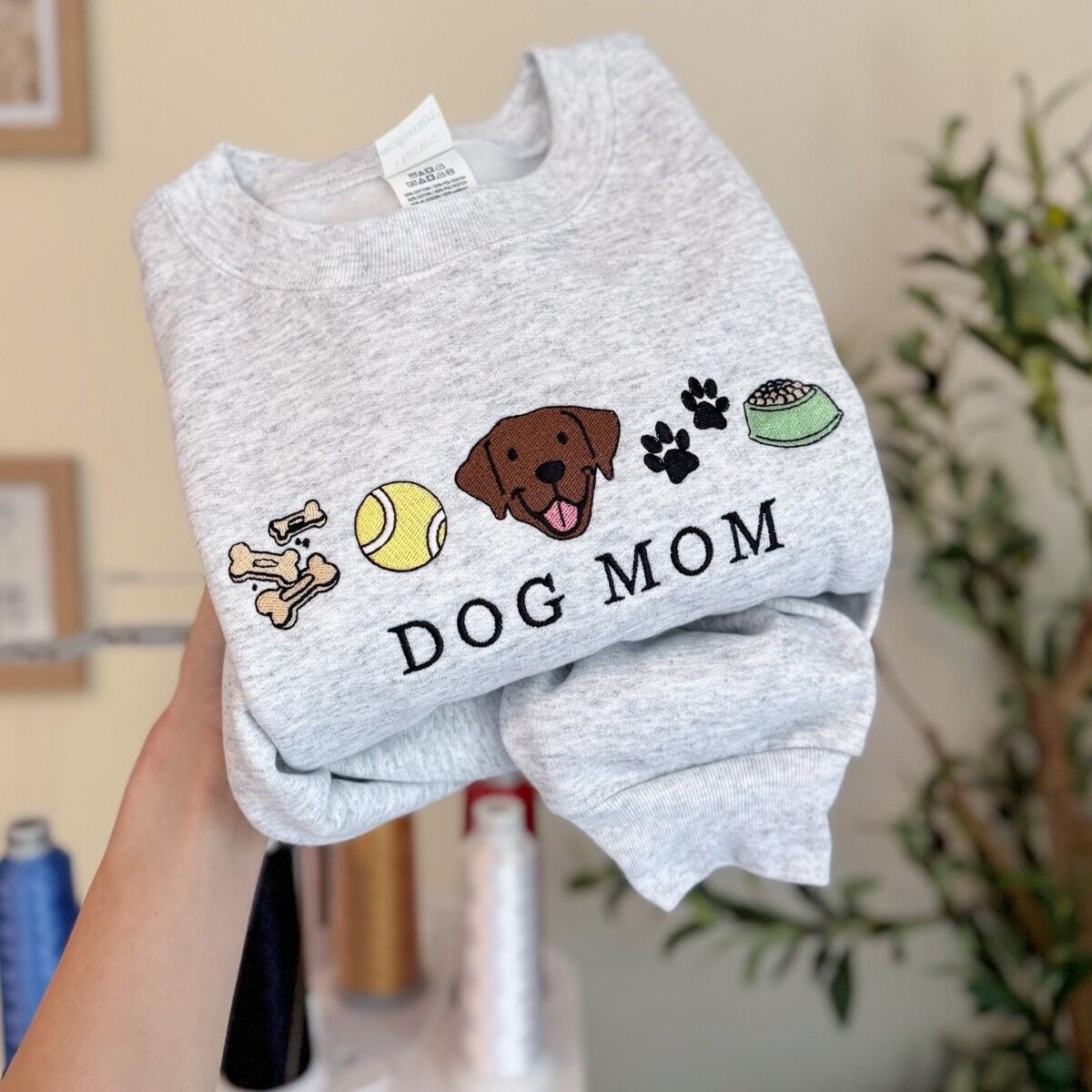 Dog Mom Icons Sweatshirt – Ash Ash crewneck sweatshirt held up in an embroidery studio, embroidered with a row of dog treats, a tennis ball, a chocolate Lab face, paw prints and a food bowl above the words DOG MOM in black thread