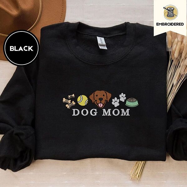 Black crewneck sweatshirt with a brown felt hat and dried bunny tail grass, embroidered with a row of dog treats, a tennis ball, a chocolate Lab face, paw prints and a food bowl above the words DOG MOM