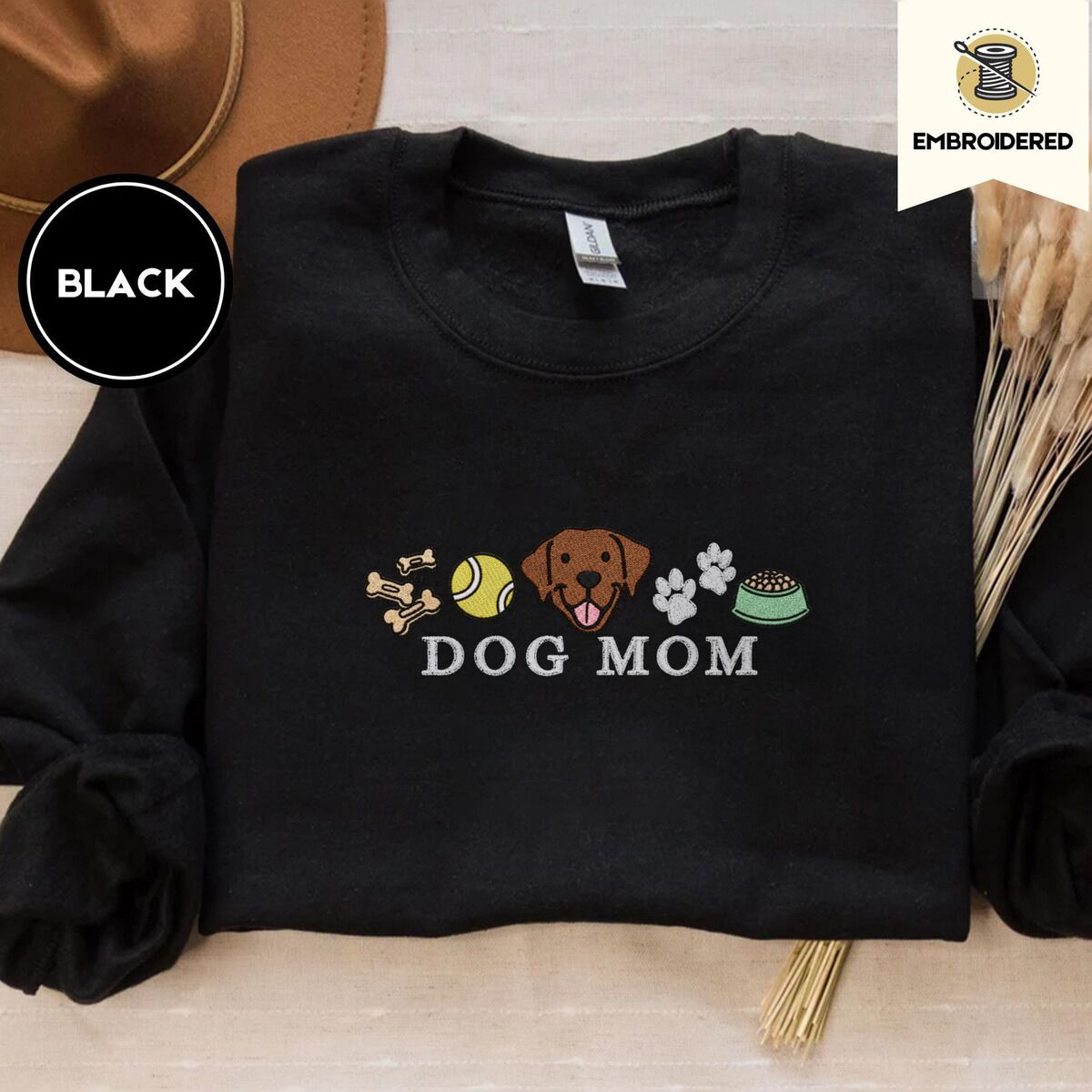 Dog Mom Icons Sweatshirt – Black Black crewneck sweatshirt with a brown felt hat and dried bunny tail grass, embroidered with a row of dog treats, a tennis ball, a chocolate Lab face, paw prints and a food bowl above the words DOG MOM