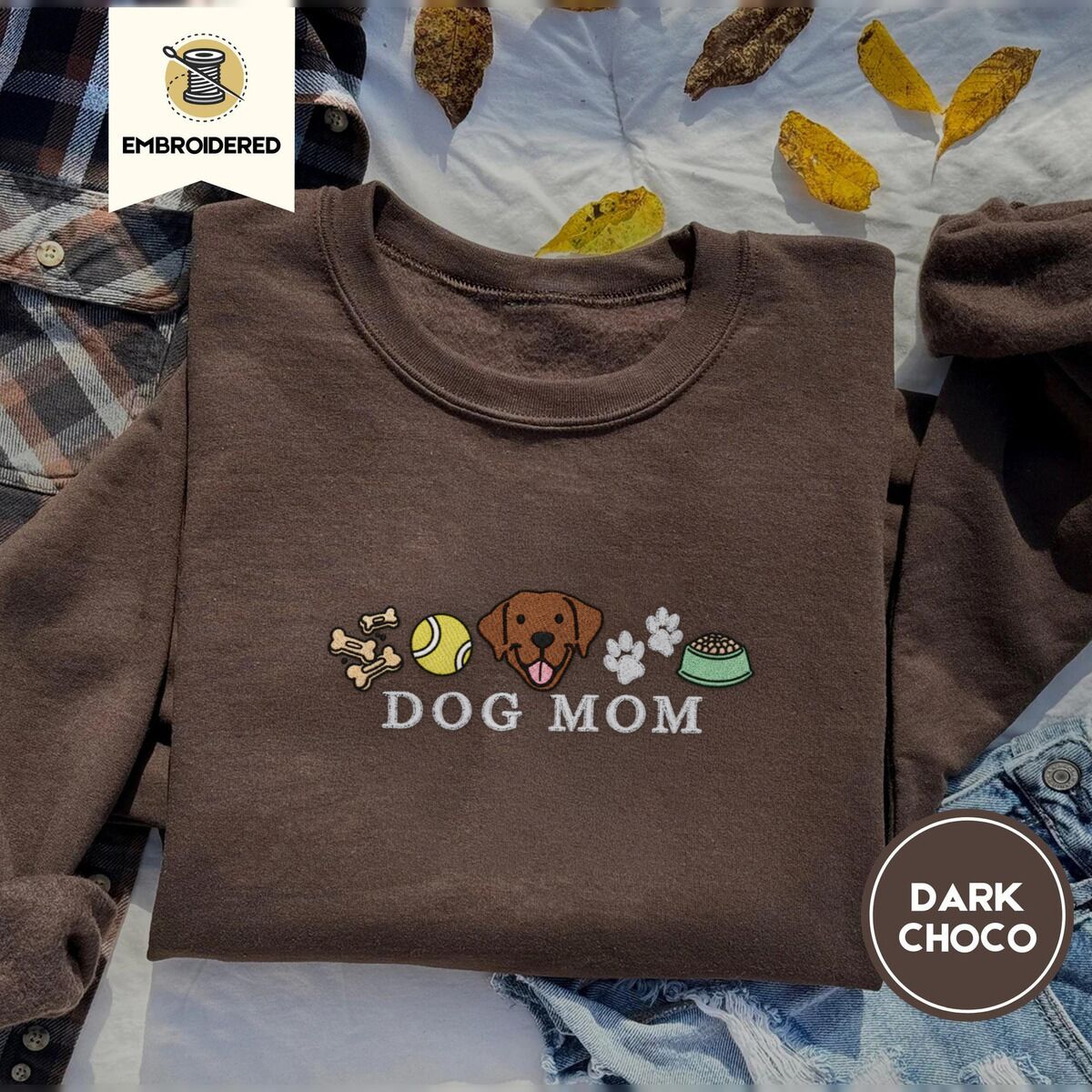 Dog Mom Icons Sweatshirt – Dark Chocolate Dark Chocolate crewneck sweatshirt on a plaid flannel shirt with yellow autumn leaves and denim shorts, embroidered with a row of dog treats, a tennis ball, a chocolate Lab face, paw prints and a food bowl above the words DOG MOM