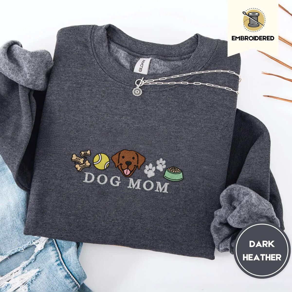 Dog Mom Icons Sweatshirt – Dark Heather Dark Heather crewneck sweatshirt folded with ripped jeans and a silver chain necklace, embroidered with a row of dog treats, a tennis ball, a chocolate Lab face, paw prints and a food bowl above the words DOG MOM