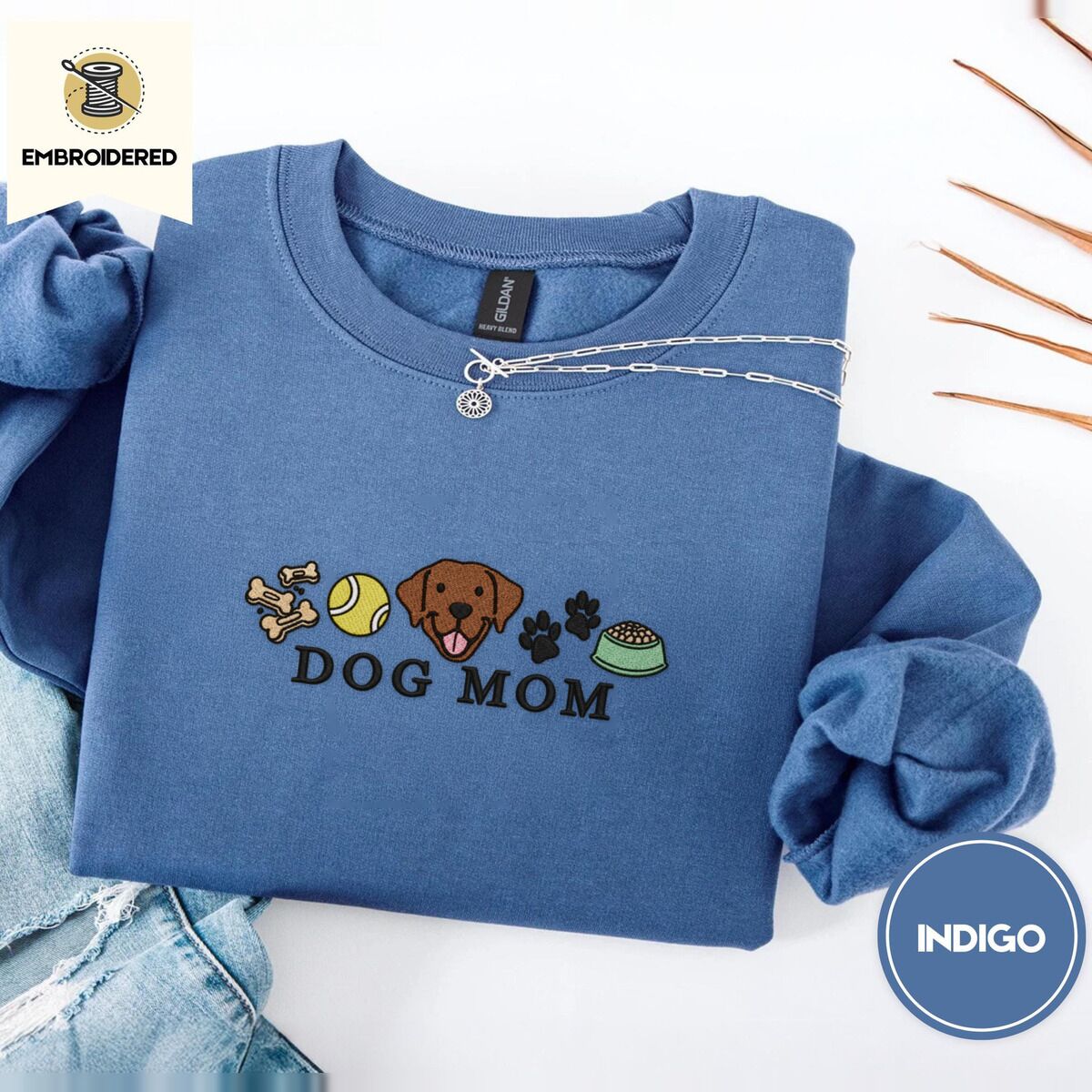 Dog Mom Icons Sweatshirt – Indigo Blue Indigo Blue crewneck sweatshirt folded with light jeans and dried twigs, embroidered with a row of dog treats, a tennis ball, a chocolate Lab face, paw prints and a food bowl above the words DOG MOM