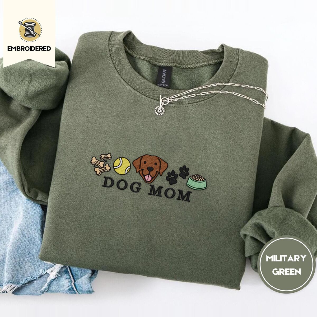 Dog Mom Icons Sweatshirt – Military Green Military Green crewneck sweatshirt folded with light jeans and a silver chain necklace, embroidered with a row of dog treats, a tennis ball, a chocolate Lab face, paw prints and a food bowl above the words DOG MOM