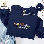 Navy crewneck sweatshirt folded with ripped jeans and a silver chain necklace, embroidered with a row of dog treats, a tennis ball, a chocolate Lab face, paw prints and a food bowl above the words DOG MOM