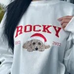 Woman wearing a white custom dog Christmas sweatshirt with ROCKY varsity letters and EST 2022